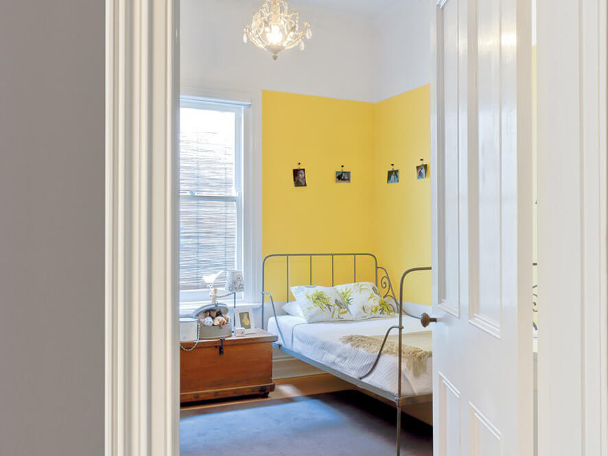 Yellow feature wall corner in bedroom with white bedding and wooden side table with photos carpet